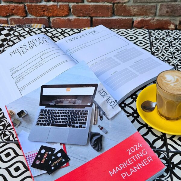 The Marketing Planner - 2024 Edition surrounded by a coffee and a book on a table next to a laptop.