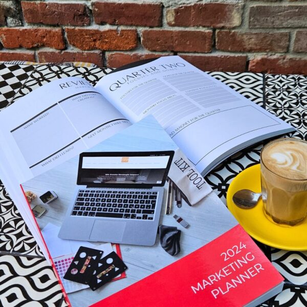 The Marketing Planner - 2024 Edition with a coffee cup and a book on a table next to a laptop.