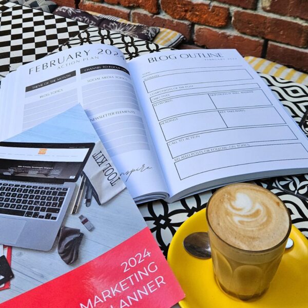 A laptop alongside a book and coffee cup placed on a table with The Marketing Planner - 2024 Edition.